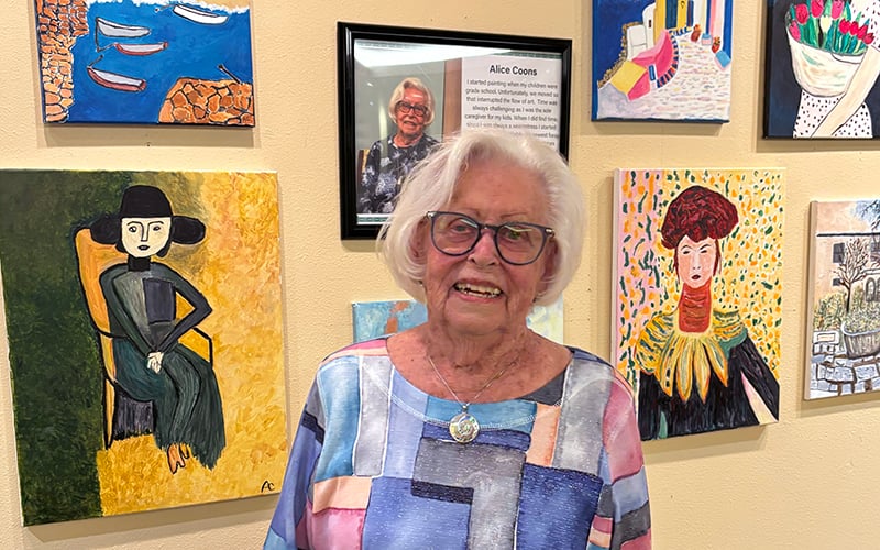 An elderly woman with white hair and glasses stands smiling in front of a display of colorful paintings and her portrait with a biography on the wall. She wears a pastel patterned blouse and a pendant necklace.