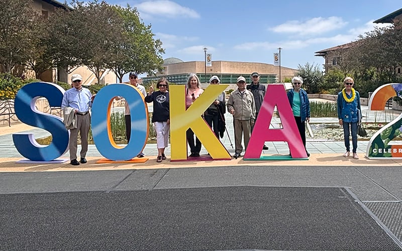 A group of people stand outdoors behind large, colorful letters spelling SOKA in front of a campus fountain and buildings on a sunny day.