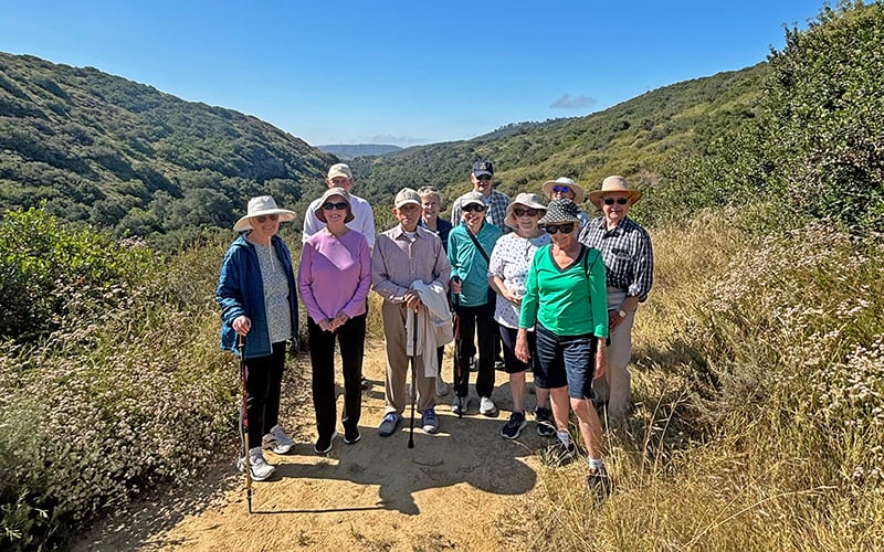 A group of older adults wearing hats and casual clothes stands on a dirt hiking trail surrounded by green hills and shrubs under a clear blue sky.
