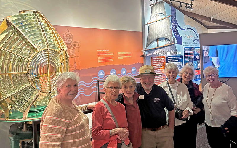 A group of seven smiling older adults stand together inside a museum. Behind them are colorful informational displays, a large glass lens, and a model sailboat on the wall. The setting has warm lighting and a welcoming atmosphere.