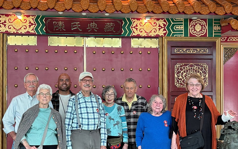 Eight smiling adults, including men and women of varying ages, stand together in front of a decorated red and gold doorway with Chinese architectural details and traditional script above the door.