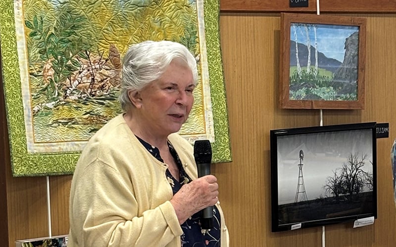An older woman with white hair and a yellow cardigan speaks into a microphone while standing in front of framed artwork displayed on a wooden wall.