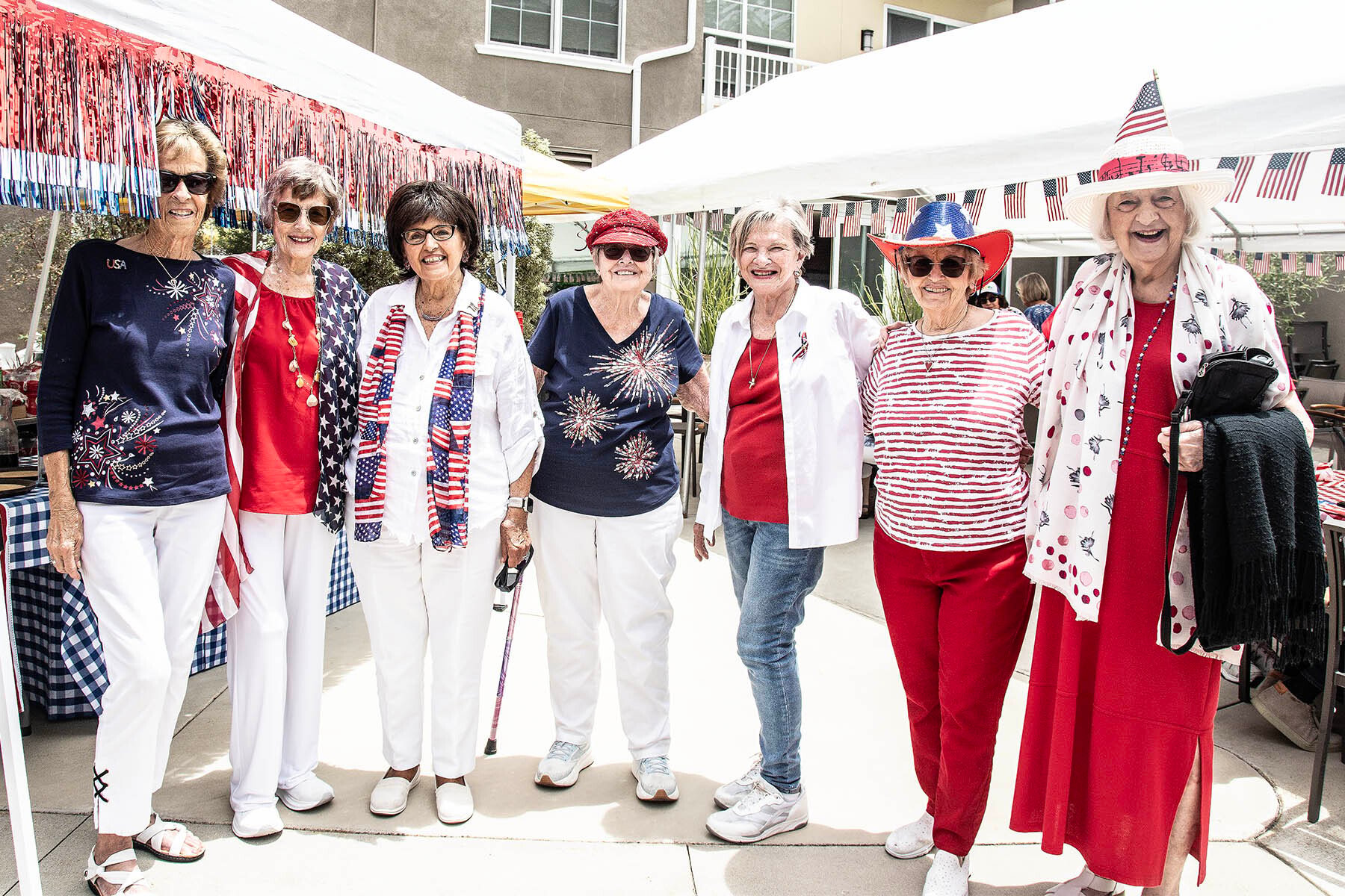 01_COV-NNC_Aug-Header Seven women stand outdoors in festive red, white, and blue outfits, smiling and celebrating, with patriotic decorations around them and a tent in the background.