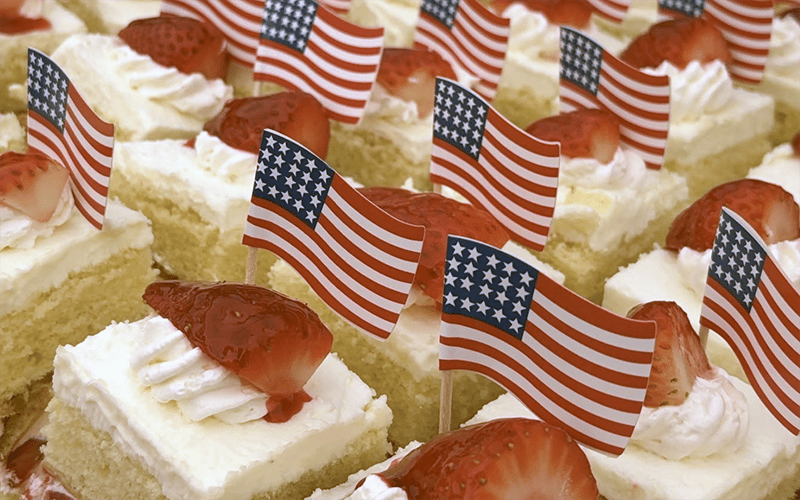 01_Cov_MemDay_2 Small square slices of cake topped with whipped cream, strawberries, and miniature American flags are arranged closely together in rows.