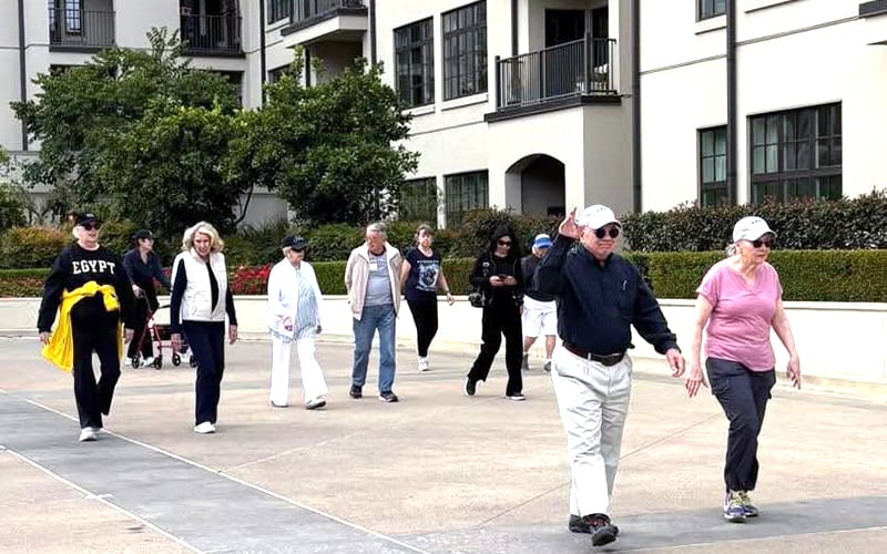 A group of people, casually dressed, are walking together outdoors on a paved path near residential buildings with balconies and greenery. Two people lead the group, smiling and appearing to enjoy the walk.
