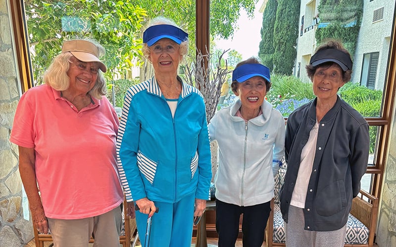 Four smiling older women stand together indoors near large windows, wearing colorful visors and casual sportswear, with greenery and a building visible outside.