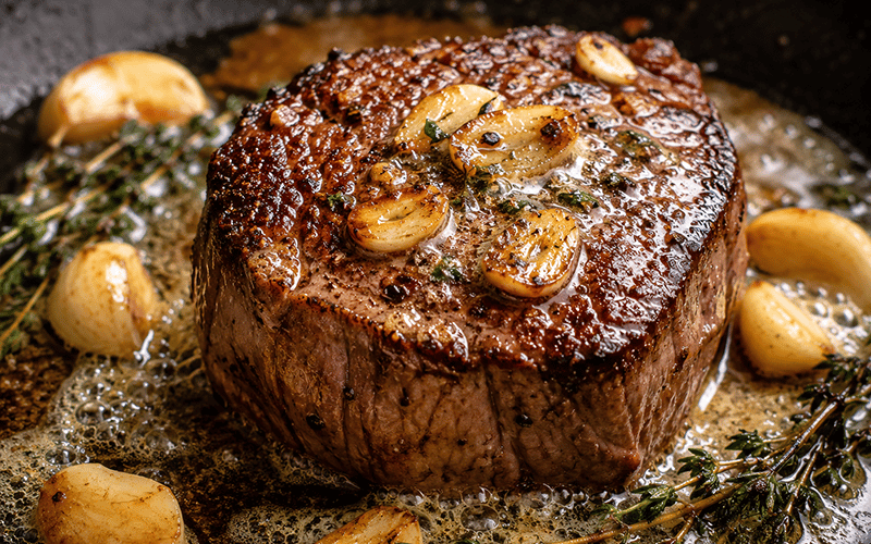 02_COV_Garlic and Butter Filet A thick, seared steak is cooking in a pan with bubbling butter, whole garlic cloves, and sprigs of fresh herbs, giving it a rich and juicy appearance.