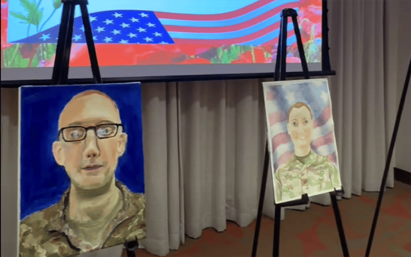 Two portrait paintings of a person in military uniform are displayed on easels. An American flag is visible in the background on a large screen above a curtained wall.