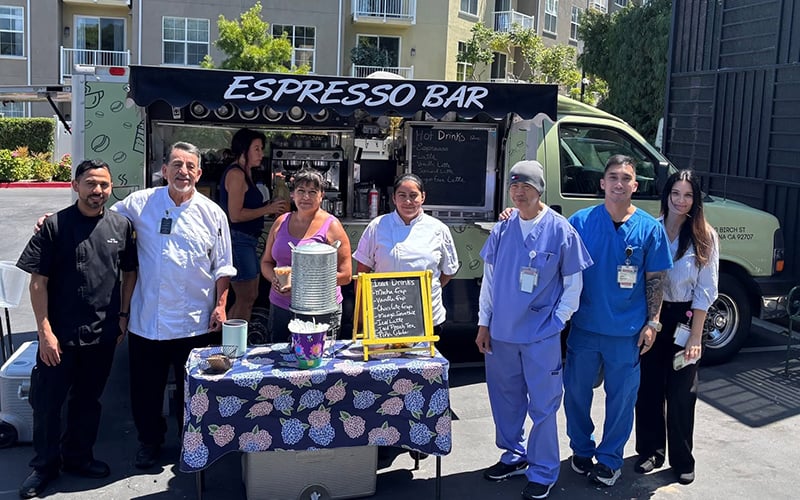 03-Story-1_COV_CoffeeBarTruck Seven people stand smiling in front of a green espresso bar truck, with a table displaying a menu and coffee supplies in the foreground. Apartment buildings and greenery are visible in the background.