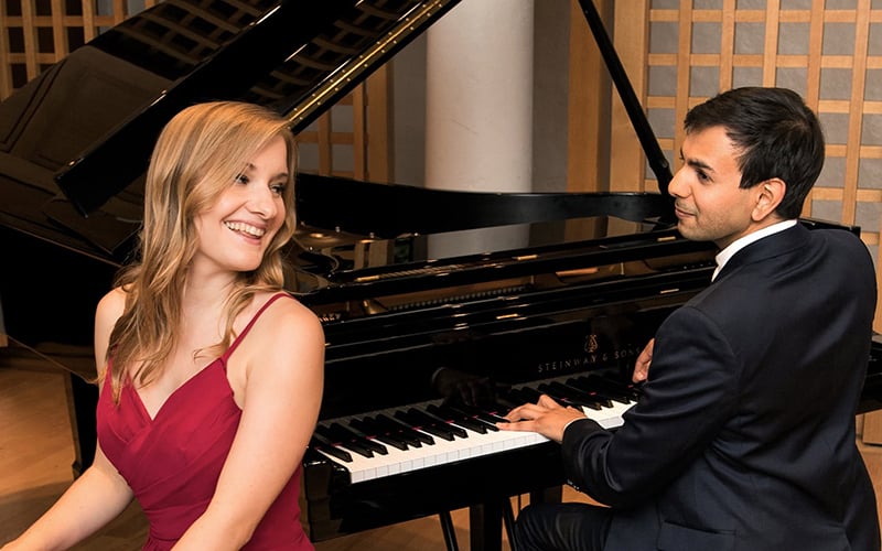 03-Story-2_COV_V Piano duo A woman in a red dress smiles while sitting beside a man in a suit who is playing a grand piano. They appear to be enjoying a musical performance together in an elegant setting.