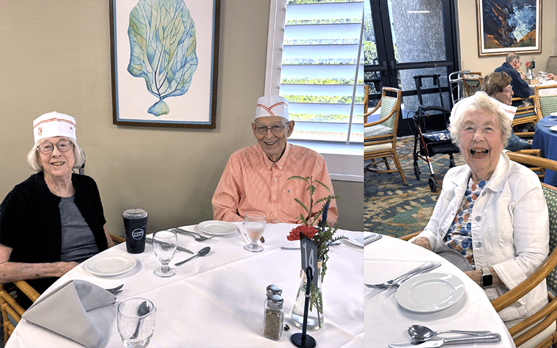 Three older adults sit smiling around a round dining table set for a meal. Two wear paper hats, and one has a large drink cup. The room is bright with art on the walls and other diners in the background.