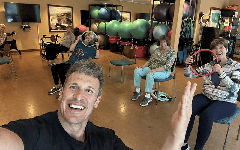 03_COV_LifeinMotion A man takes a selfie with a group of smiling older adults in a fitness studio. They are seated on chairs, holding exercise equipment, with stability balls and fitness gear visible in the background.
