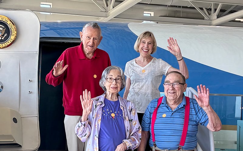 Four older adults, three standing and one seated, smile and wave at the camera in front of a plane with the presidential seal visible on the door. They are wearing casual clothes and name tags, inside a brightly lit hangar.