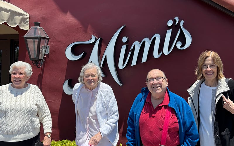 Four smiling seniors, three women and one man, stand together in front of a red wall with a large Mimis sign, enjoying a sunny day.