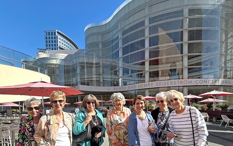 05_COV_Sound of Music A group of eight older women stand smiling together outdoors in front of the modern glass facade of Renee and Henry Segerstrom Concert Hall on a sunny day. Pink umbrellas and tables are in the background.
