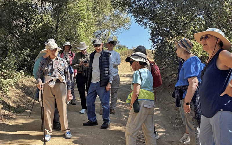 06_COV_Laguna Canyon A group of people, mostly older adults, stand and chat on a sunlit dirt trail surrounded by trees. Many wear hats, sunglasses, and casual outdoor clothing; one person uses a walking stick.