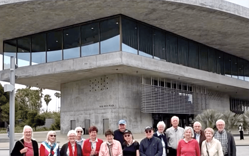 A group of older adults stands smiling in front of a modern concrete building with large glass windows and overhanging roof. Palm trees and greenery are visible in the background.