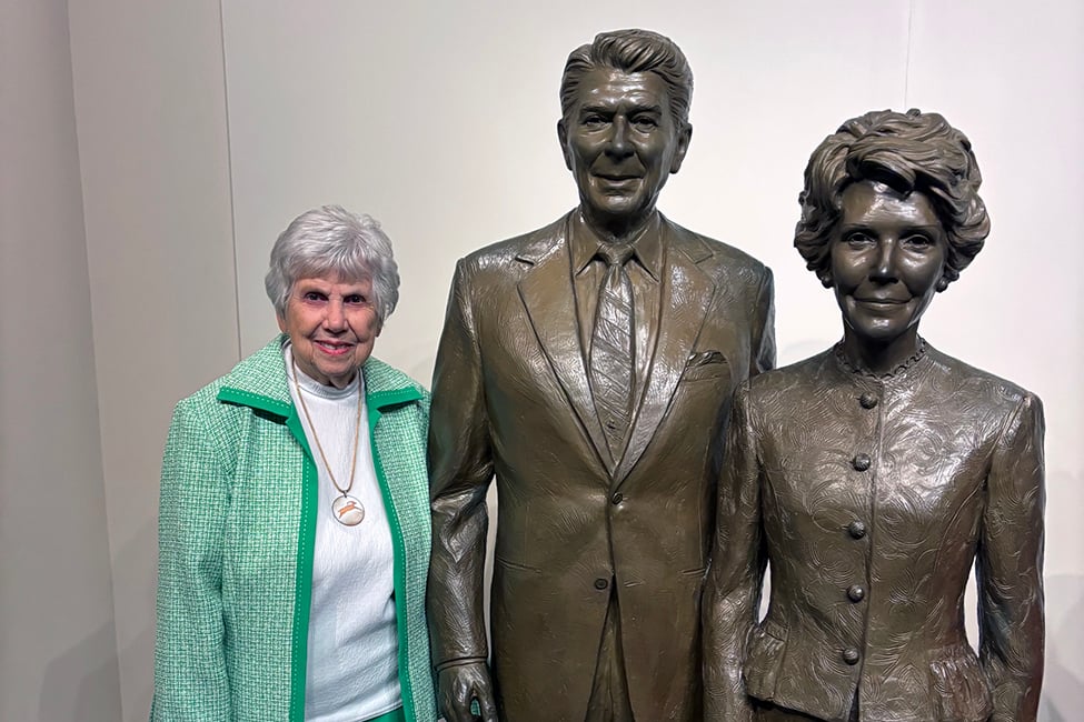 An older woman with short gray hair stands smiling next to bronze statues of a man in a suit and a woman in a buttoned jacket, against a plain background.