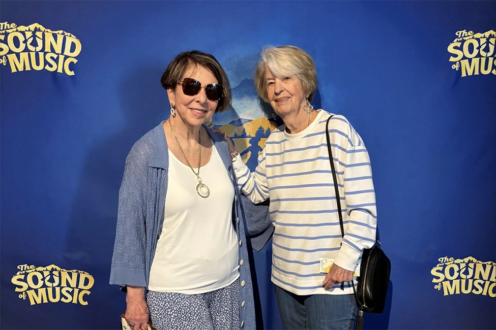 Cov_SofM_Header Two older women stand together smiling in front of a blue The Sound of Music backdrop. One wears sunglasses, a blue cardigan, and necklace; the other wears a striped top, holding a bag over her shoulder.