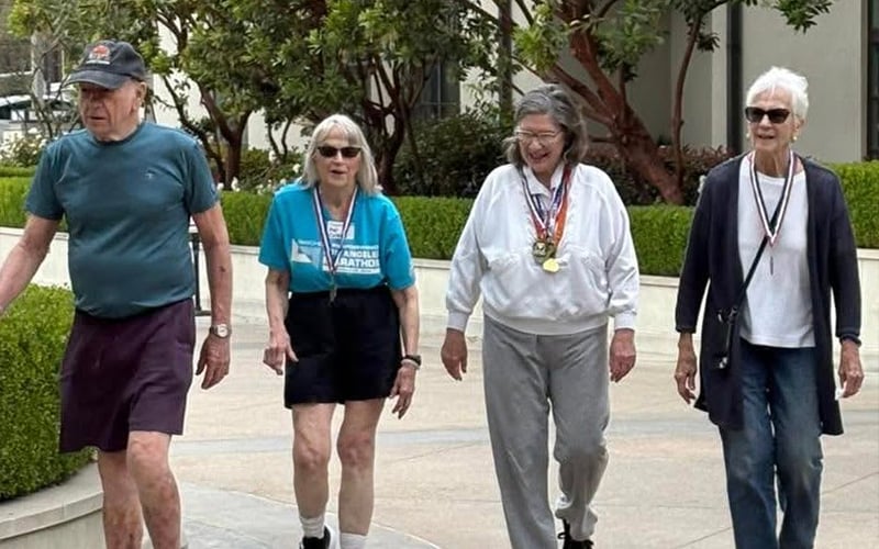 Four older adults walk outdoors on a paved path. Three women and one man are smiling; some wear medals around their necks. Trees and bushes are in the background, suggesting a pleasant, sunny day.