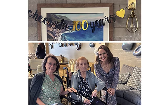 Three women sit and stand together, smiling, in front of a cheers to 100 years banner at a celebration. One woman is in a wheelchair, and the group is surrounded by festive decorations.