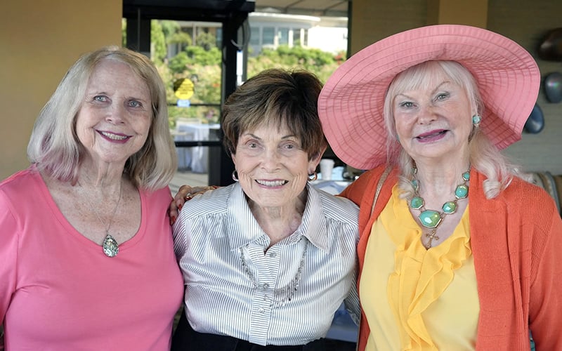 Three smiling older women pose together indoors. One wears a pink top, another a striped blouse, and the third a pink hat and yellow blouse with a turquoise necklace.