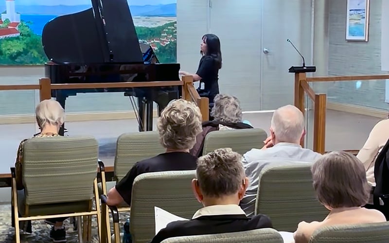 A woman plays a black grand piano in front of an audience of older adults seated in rows, some reading from papers. A large painting and a microphone are visible in the background.