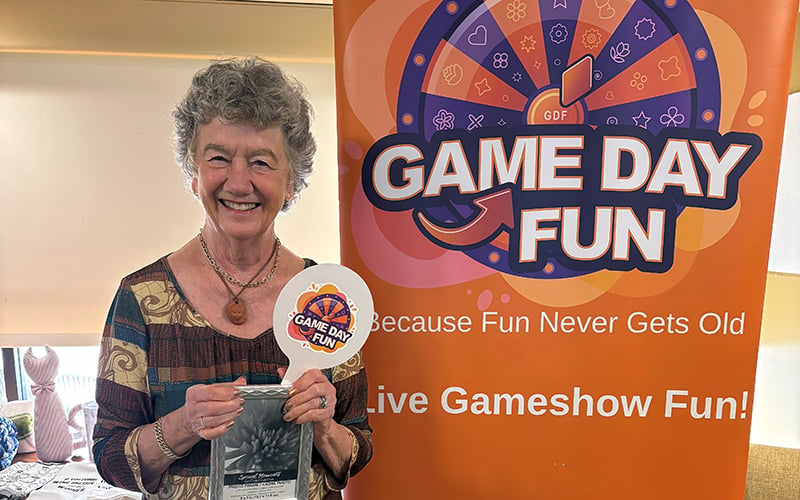 An older woman smiles while holding a Game Day Fun paddle in front of an orange Game Day Fun banner that says, Because Fun Never Gets Old. Live Gameshow Fun!.