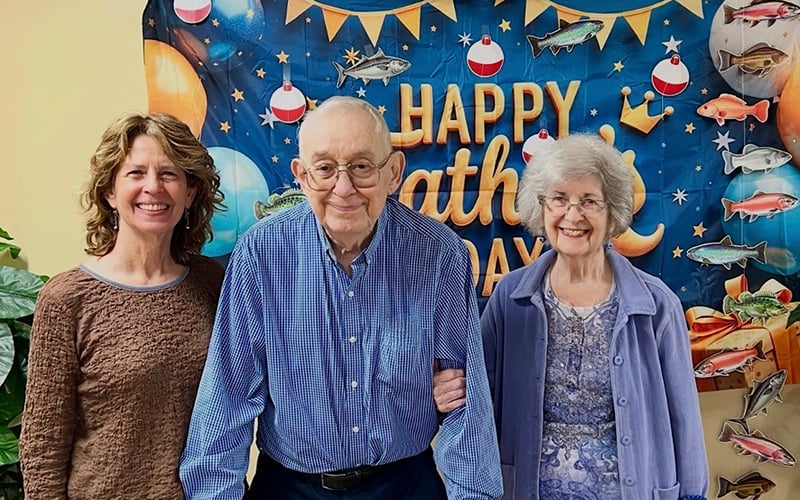 Three people stand smiling in front of a Happy Fathers Day banner decorated with stars, moons, and fish. The group includes an older man between two women. Green plants are partially visible on the left.