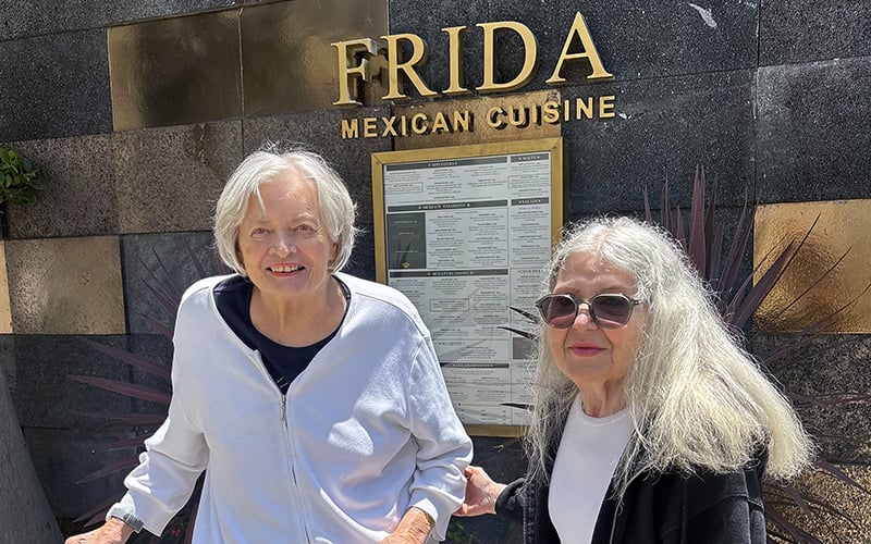 Two older women stand smiling in front of a FRIDA Mexican Cuisine restaurant sign and menu. One wears a white jacket, the other sunglasses and a dark top. Sunlight shines on them.