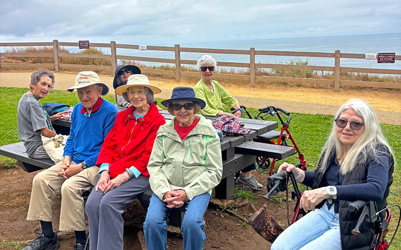 Seven older adults sit and smile on benches by the ocean, some wearing hats and sunglasses. A walker and cane are visible. It is partly cloudy, with grassy and sandy areas near a wooden fence overlooking the water.