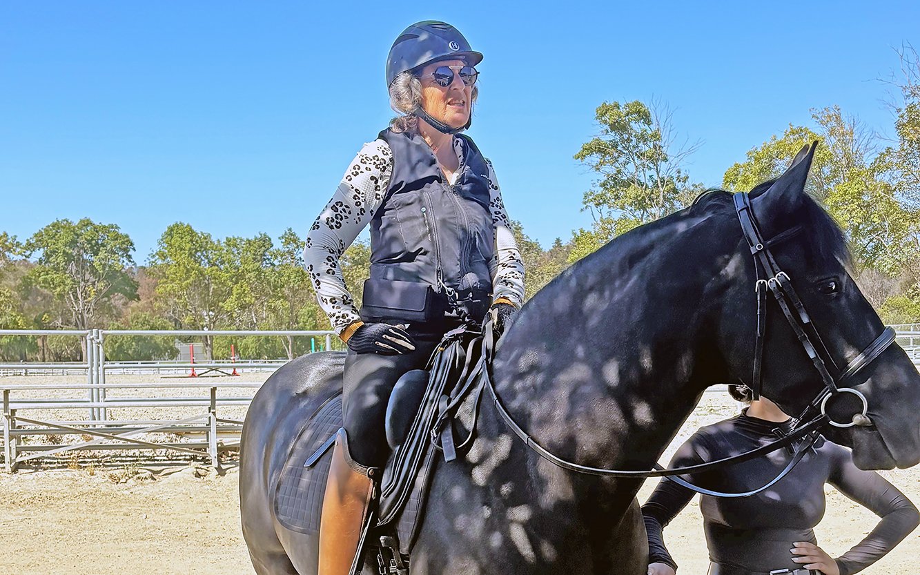 An older woman wearing a helmet, sunglasses, and riding gear sits confidently on a black horse outdoors, with trees and blue sky in the background.