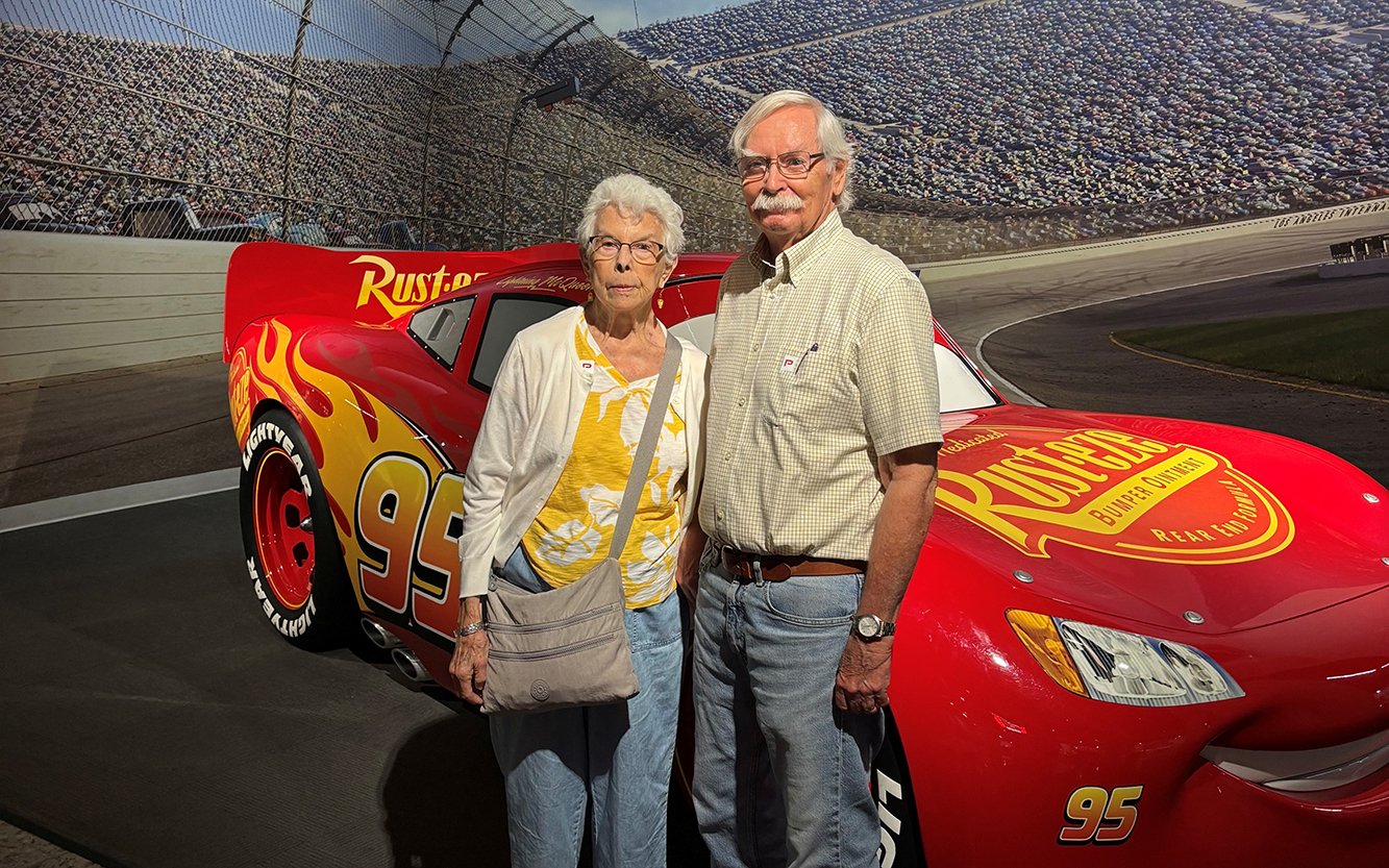 An older couple stands smiling in front of a red animated race car display with the number 95, inside a building that looks like a racetrack.