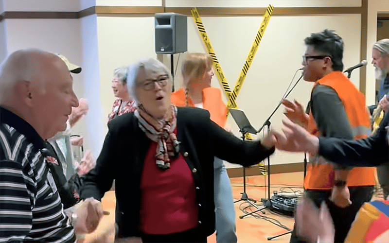 03_Story-2_MC_Aud Closing Prty_dancing Older adults and younger people wearing orange vests dance and interact energetically indoors, with yellow caution tape and music equipment visible in the background.