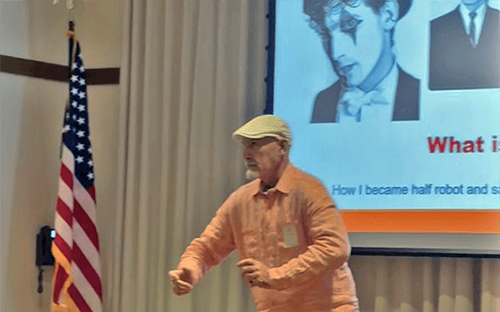 03_Story-3_MC_Zoot A man in a light cap and peach shirt gestures in front of a U.S. flag and a projection screen showing a black-and-white photo of a person in makeup. The room has light curtains and a presentation is in progress.