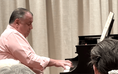03_Story-4_MC_Conservatory_Pianist A man in a pink shirt plays a grand piano on stage, with sheet music open in front of him. An audience watches, partially visible in the foreground.