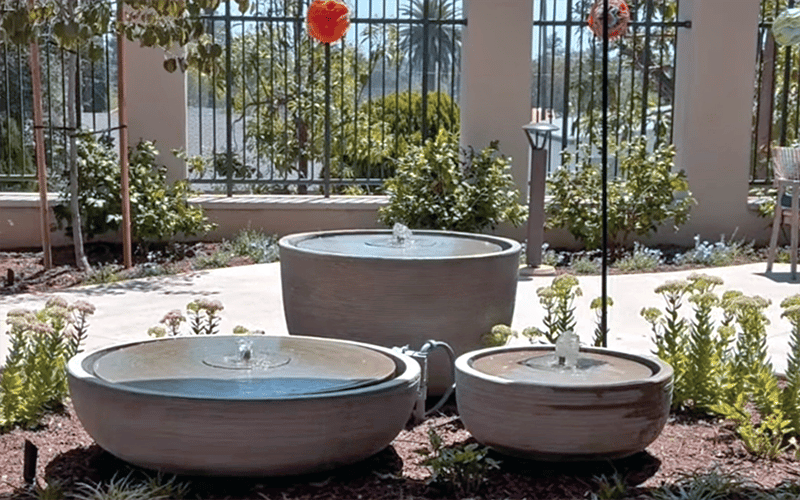 03_Story-5_MC_Memory care patio opening Three round, tiered garden fountains of varying heights sit among flowers and greenery, with water gently bubbling from each. A fence and trees are visible in the background under a sunny sky.