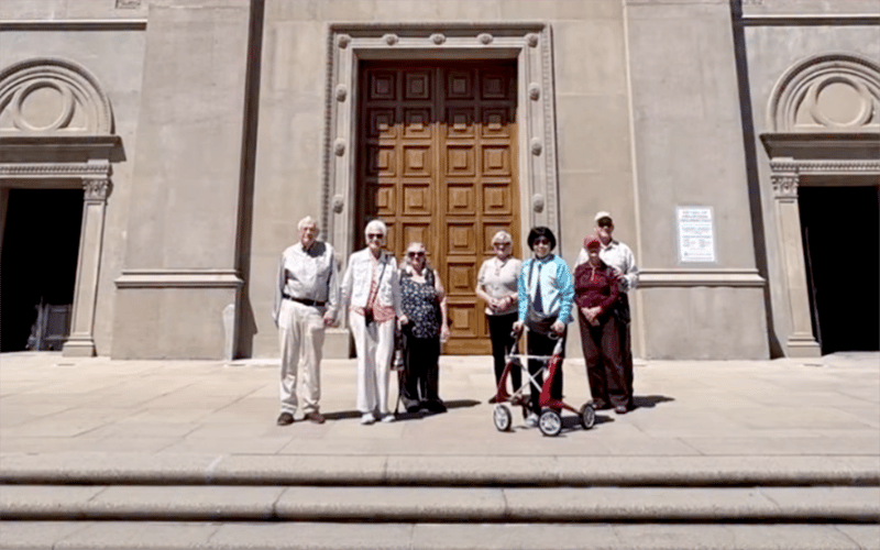 03_Story-6_MC_Forest Lawn trip Seven older adults stand on stone steps in front of a large wooden door on a stone building. One person uses a walker. It appears to be sunny.