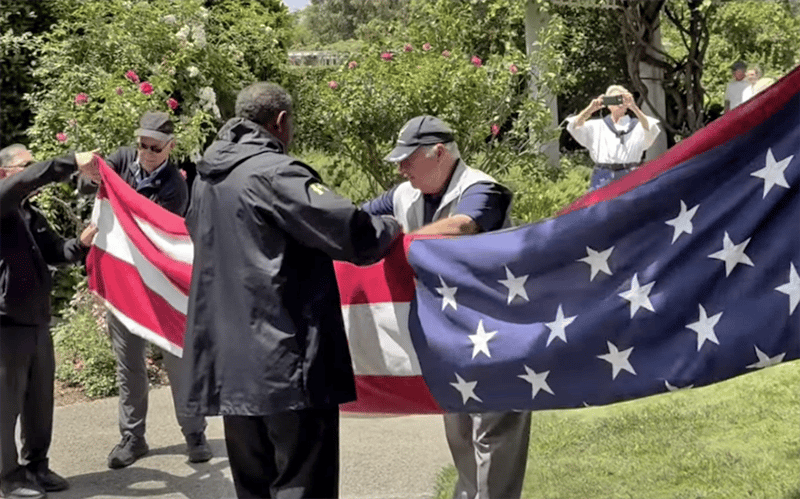 03_Story-7_MC_Flag donation to Huntington Four people hold and fold an American flag outside, while another person in the background takes a photo. There is greenery and flowers around them.