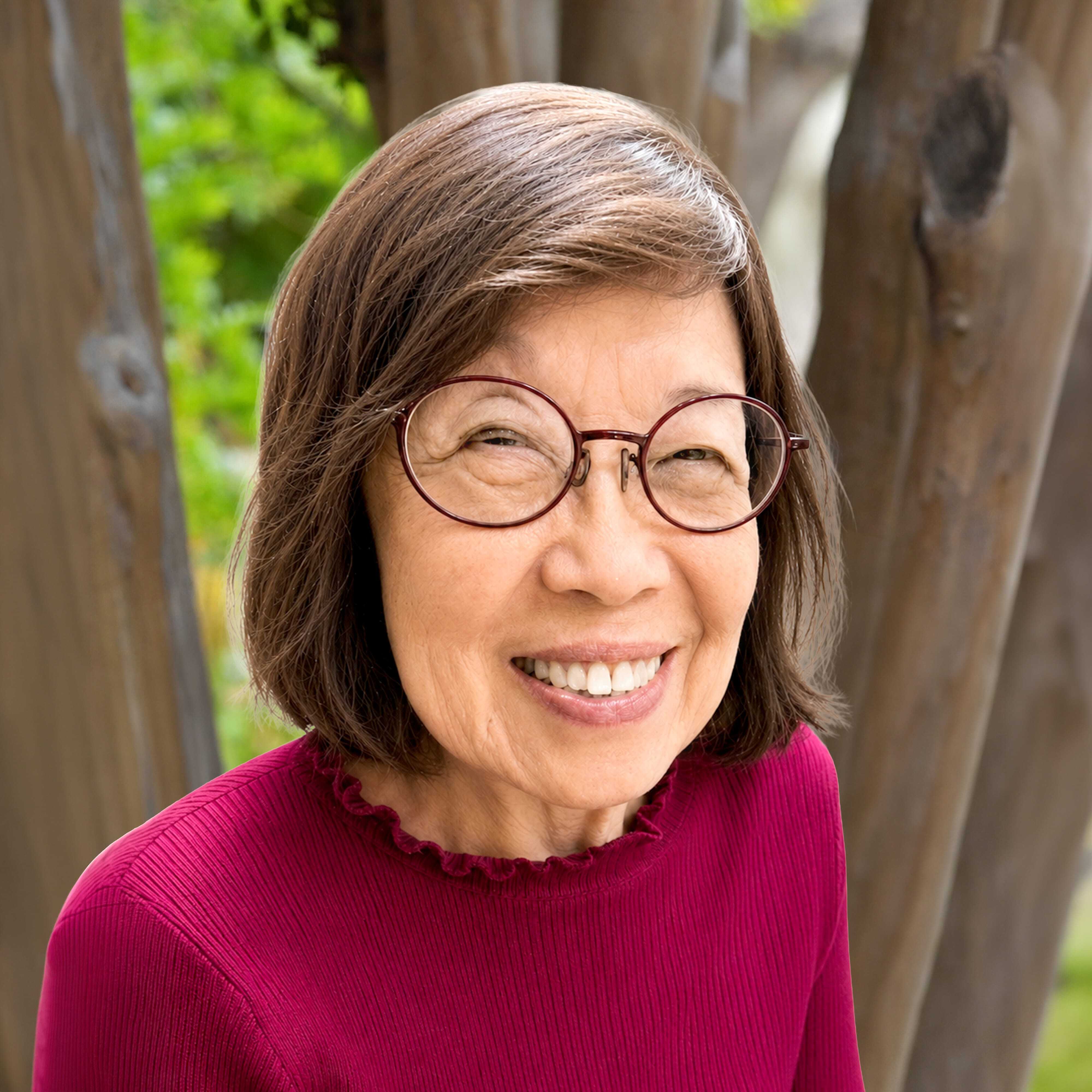 An older woman with short brown hair, wearing round glasses and a red top, smiles outdoors in front of a tree with green foliage in the background.