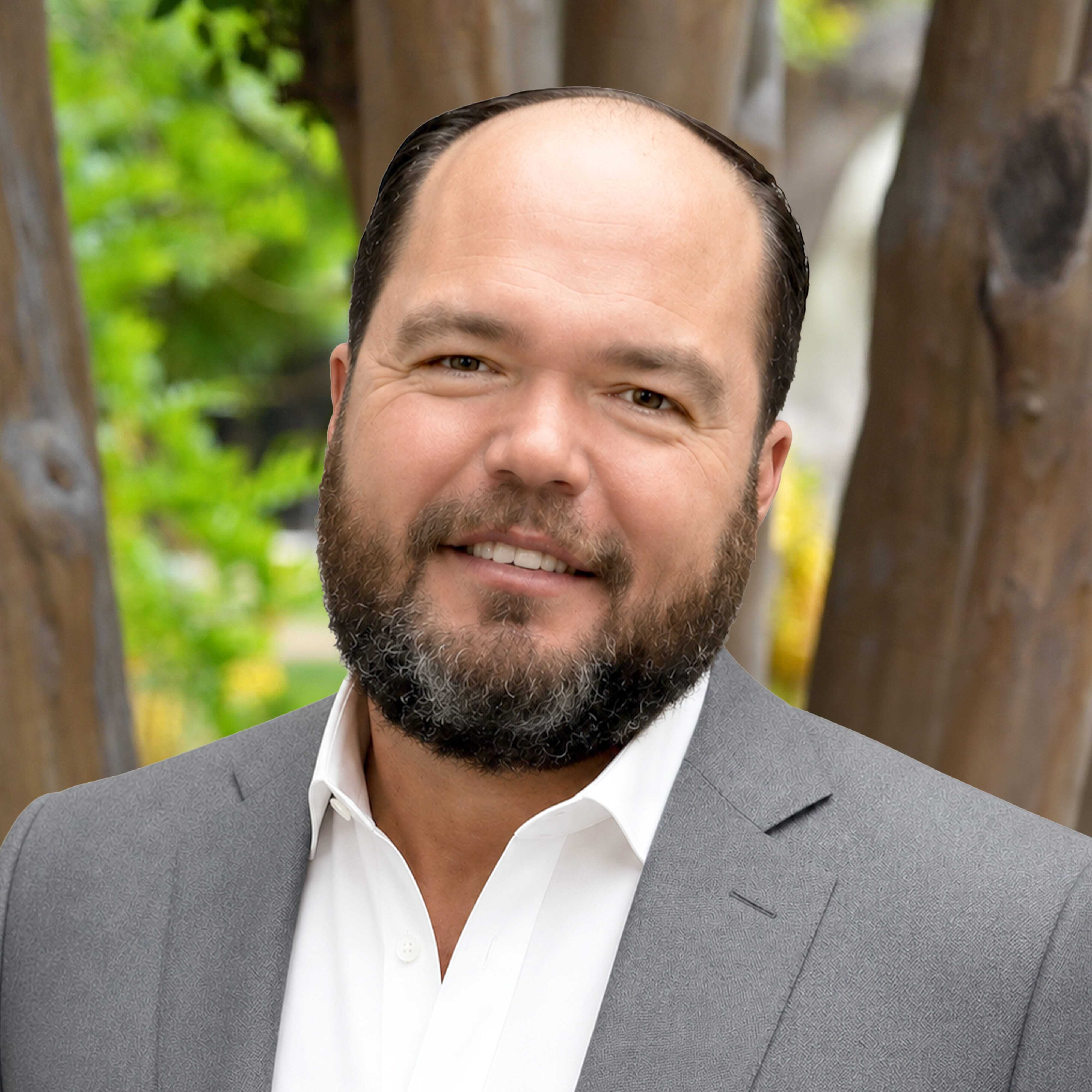 Smiling man with a beard and mustache wearing a gray suit jacket and white shirt, standing outdoors in front of green foliage and tree trunks.