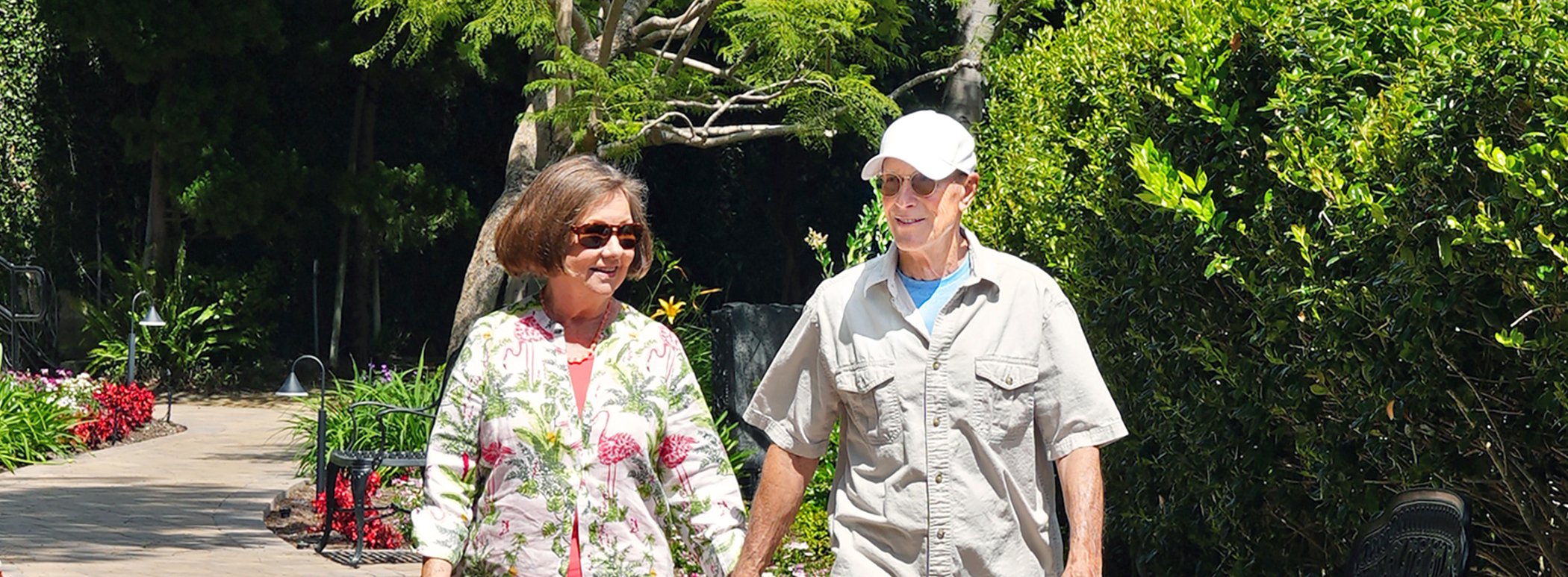 Older couple holding hands and walking outside on a sunny day, surrounded by lush greenery and flowers along a paved path.
