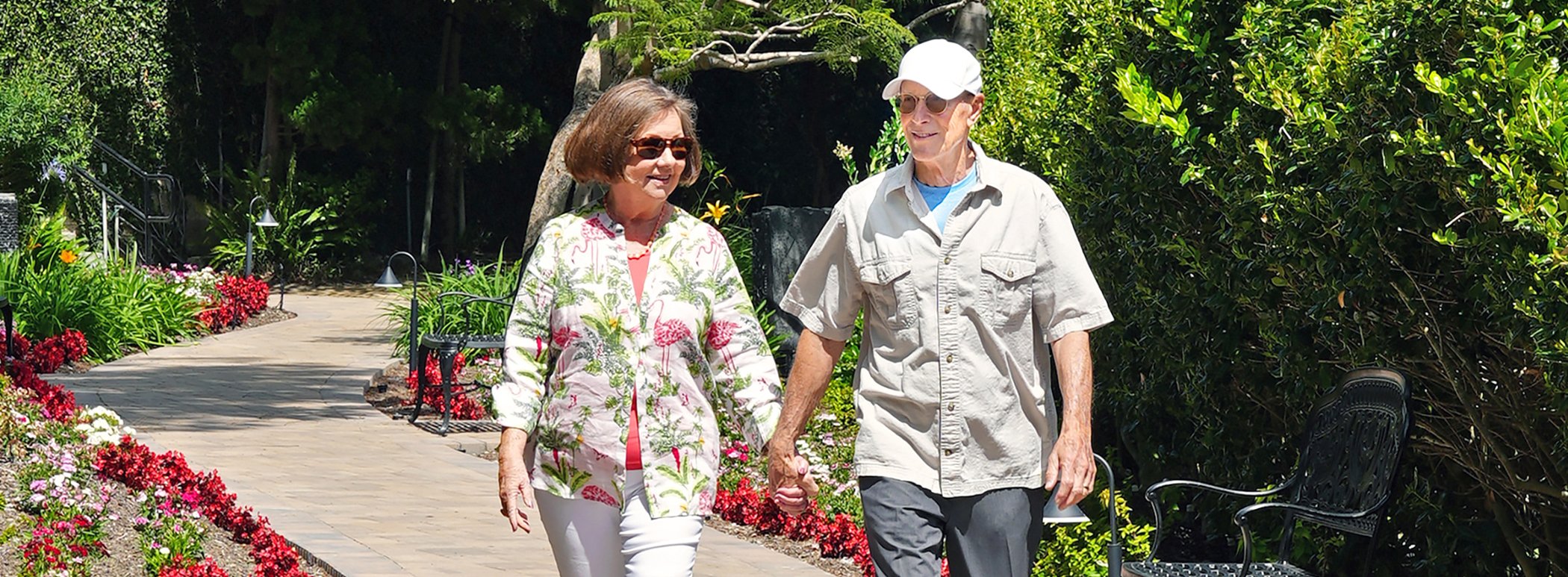 An older couple holding hands walks along a sunny garden pathway lined with flowers, greenery, and benches.