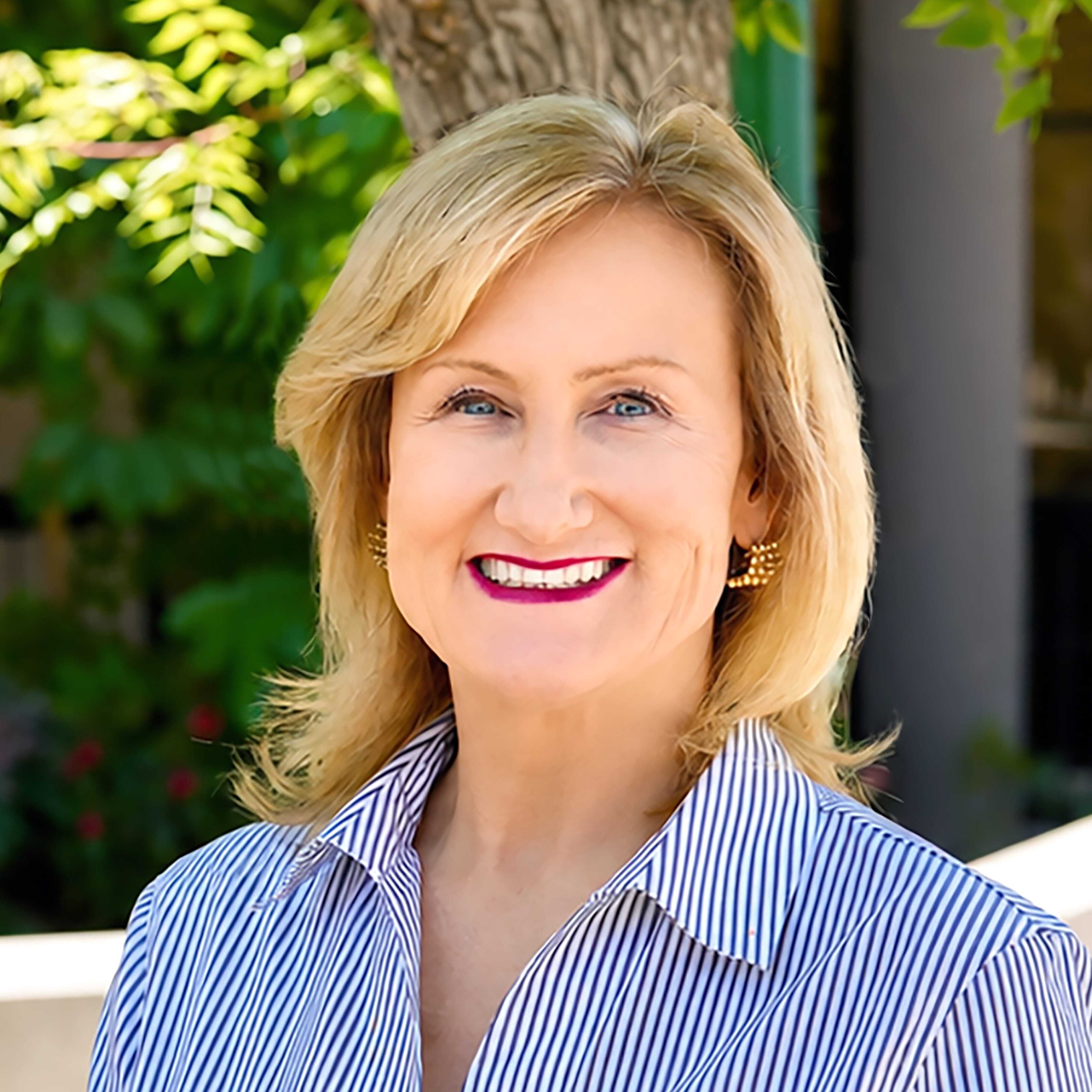 A woman with blonde hair, wearing a striped button-up shirt and gold earrings, smiles outdoors with trees and greenery in the background.