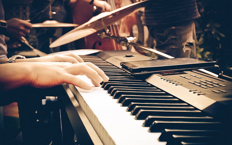 Close-up of hands playing a digital keyboard, with a smartphone resting on the keyboard. Drums and other musicians are visible in the blurred background. The scene has a warm, ambient lighting.