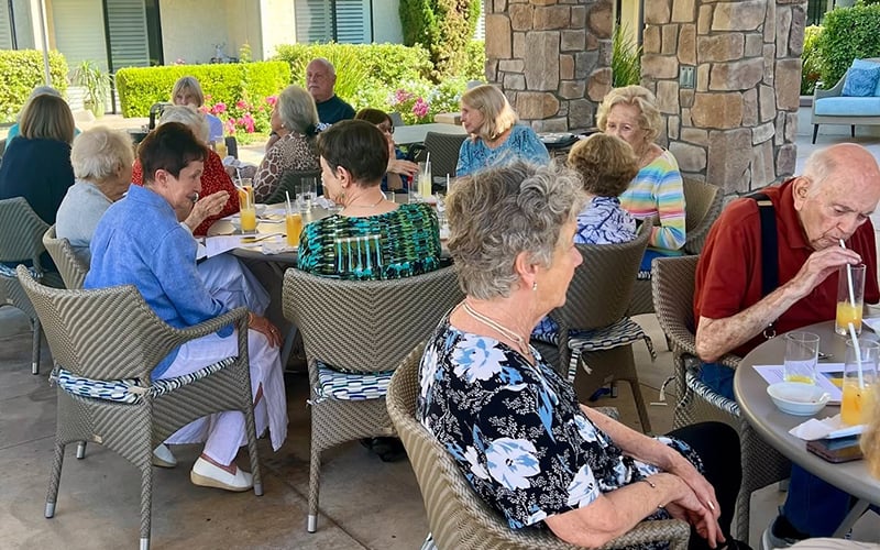 01_CAN-Header_Sept2026.png A group of seniors sit outside at tables, eating and drinking together in a garden patio area, enjoying a sunny day and socializing.
