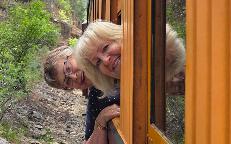 Two older women smiling and leaning out of the window of a yellow train as it travels through a green, wooded area.
