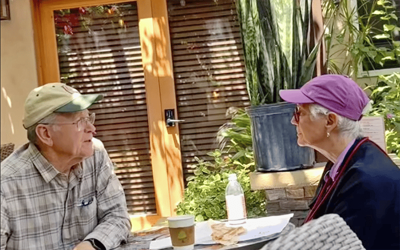 01_MC-Header_Sept2026 An older man and woman sit at an outdoor table, talking. There is coffee, a water bottle, and food on the table. Plants and a door are in the background.