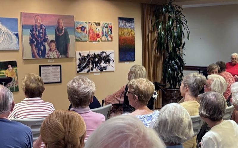 A group of elderly people sit facing a wall displaying colorful paintings and art pieces at what appears to be an art exhibit or community event.