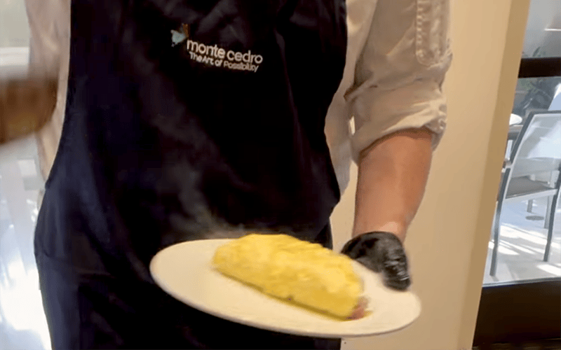 03-Story_01_ChefDemo A person wearing a monte cedro apron holds a white plate with a yellow omelette, standing indoors near a window.